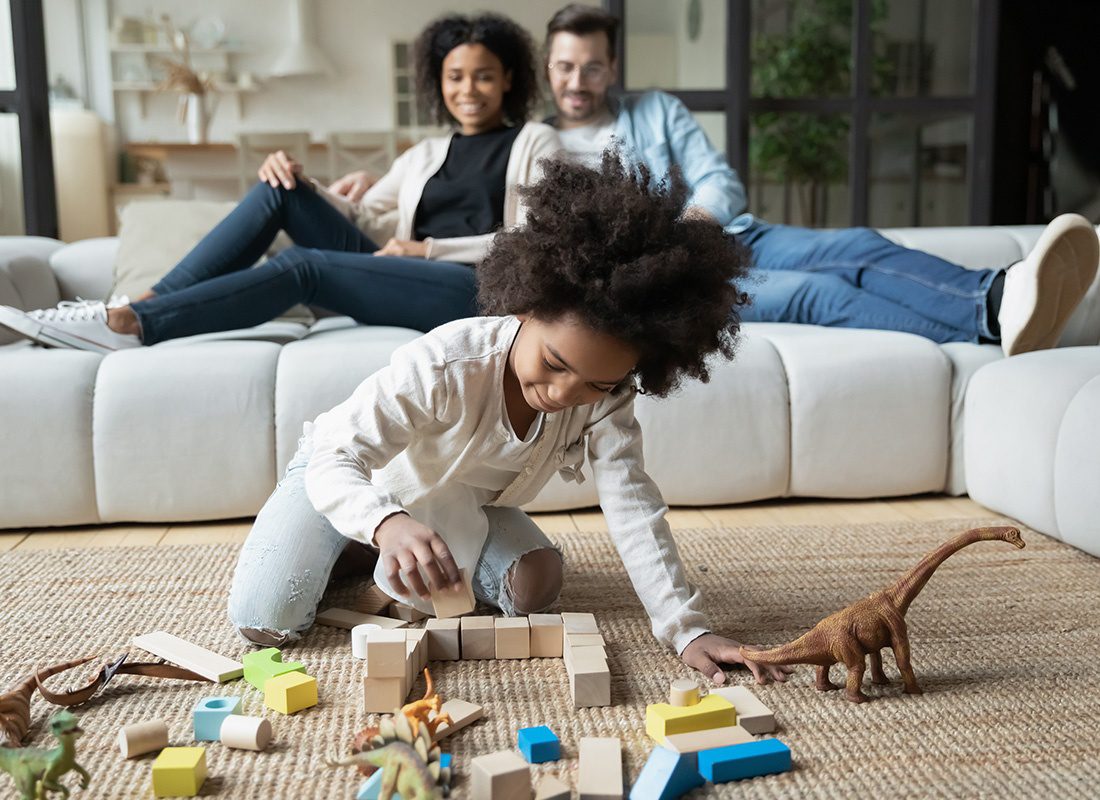 Personal Insurance - Happy Mother and Father Sit on a Sofa as They Watch Their Daughter Play With Blocks and Toy Dinosaurs